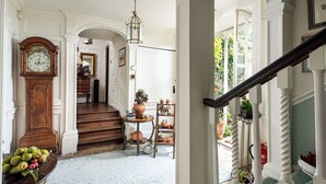 A grand hallway with a wooden staircase, a large grandfather clock, and a variety of potted plants. - Compton House (Axbridge)