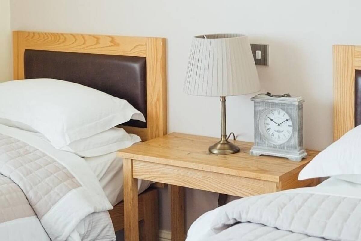 A hotel room with a wooden headboard, a bedside table with a lamp and a clock, and two beds with white bedding.