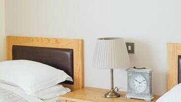 A hotel room with a wooden headboard, a bedside table with a lamp and a clock, and two beds with white bedding.