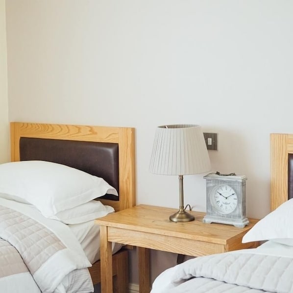 A hotel room with a wooden headboard, a bedside table with a lamp and a clock, and two beds with white bedding. - The Jolly Drover (Liss)
