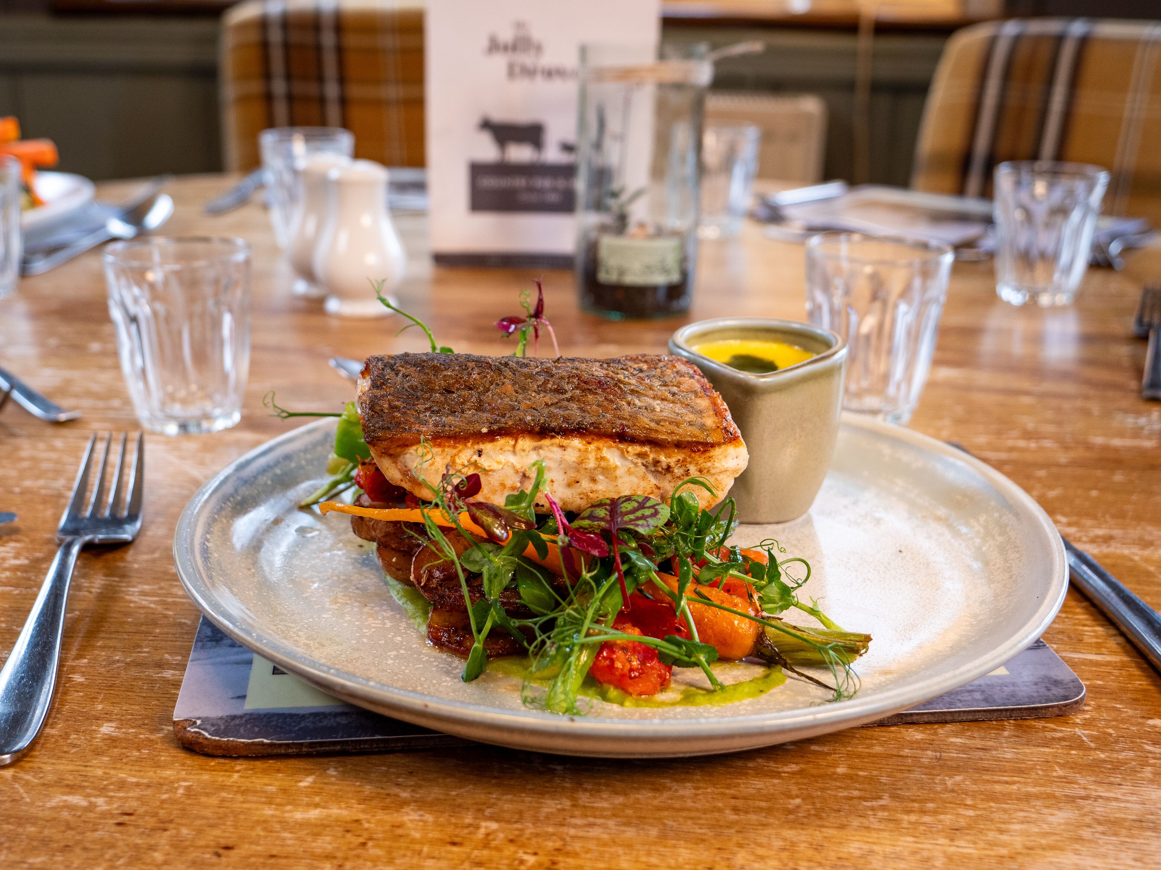 Un plato con un sándwich de pescado a la plancha, un cuenco pequeño de salsa y una variedad de hojas verdes y vegetales.