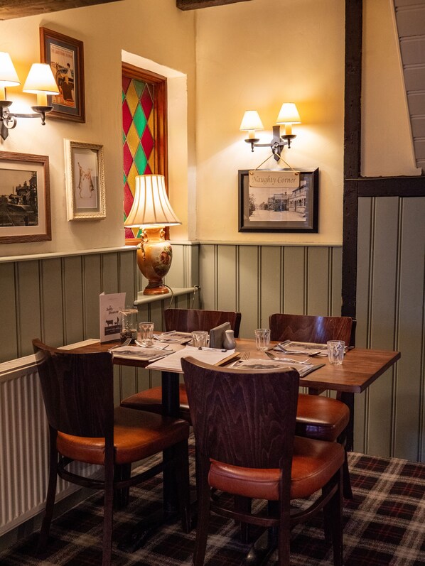 A dining area with a table set for four, surrounded by wooden chairs. There are framed pictures on the wall, a colorful diamond-patterned window, and wall-mounted lamps.
