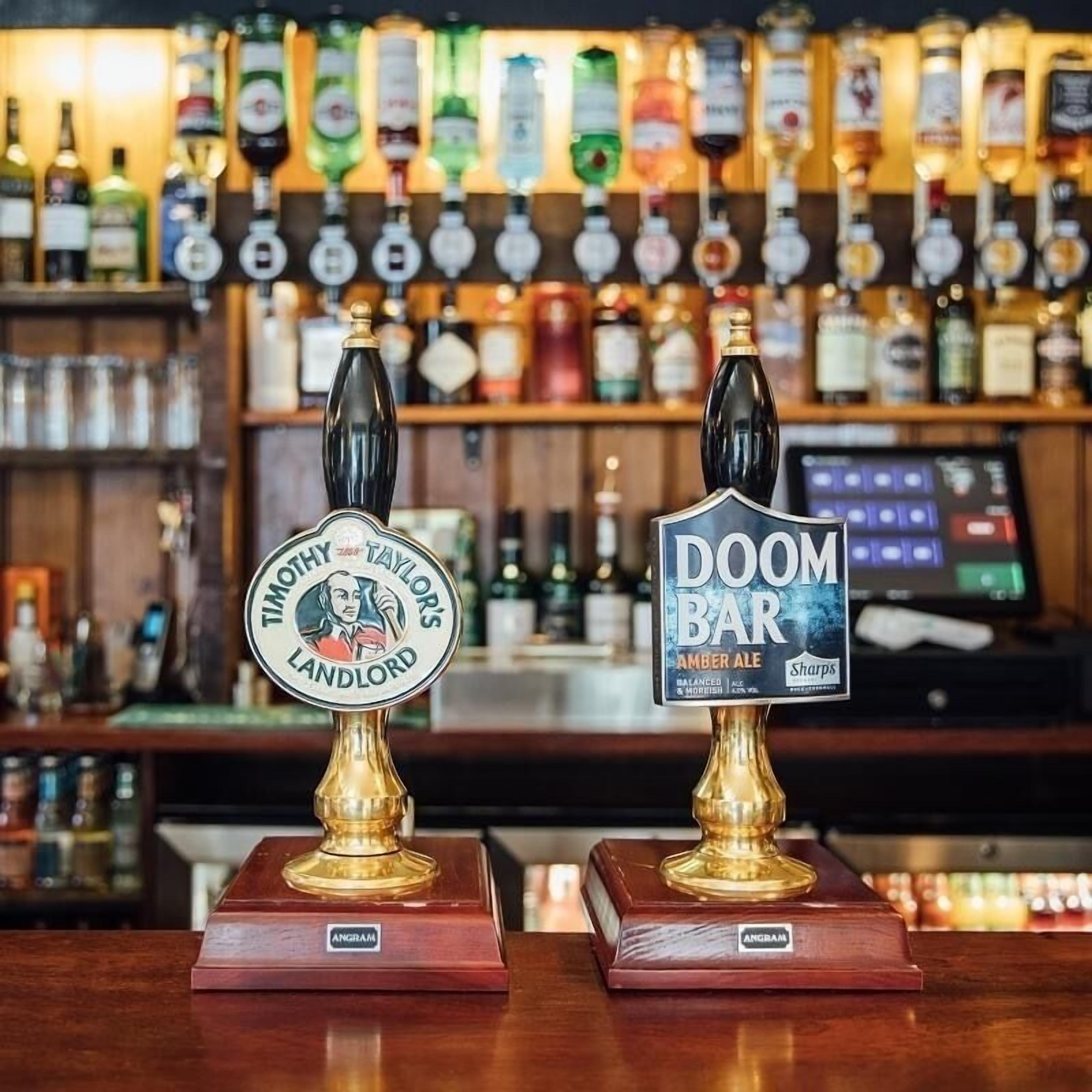 Dos grifos con etiquetas, uno que dice 'MOTLEY TAYLOR'S LANDLORD' y otro 'DOOM BAR AMBER ALE', sobre una barra con un fondo de varias botellas.