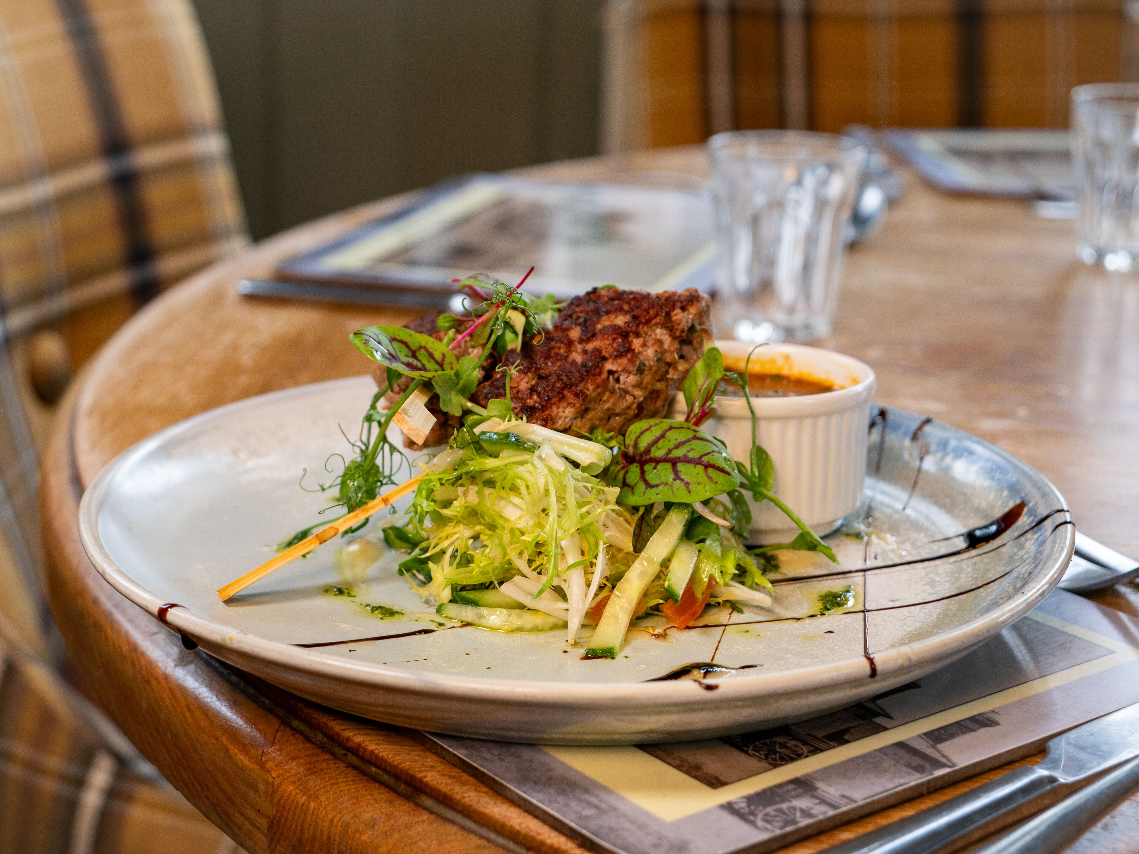 A plate of food with a salad and a meat dish, accompanied by a small bowl of sauce, on a wooden table with a placemat.