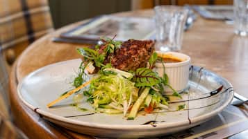 A plate of food with a salad and a meat dish, accompanied by a small bowl of sauce, on a wooden table with a placemat.