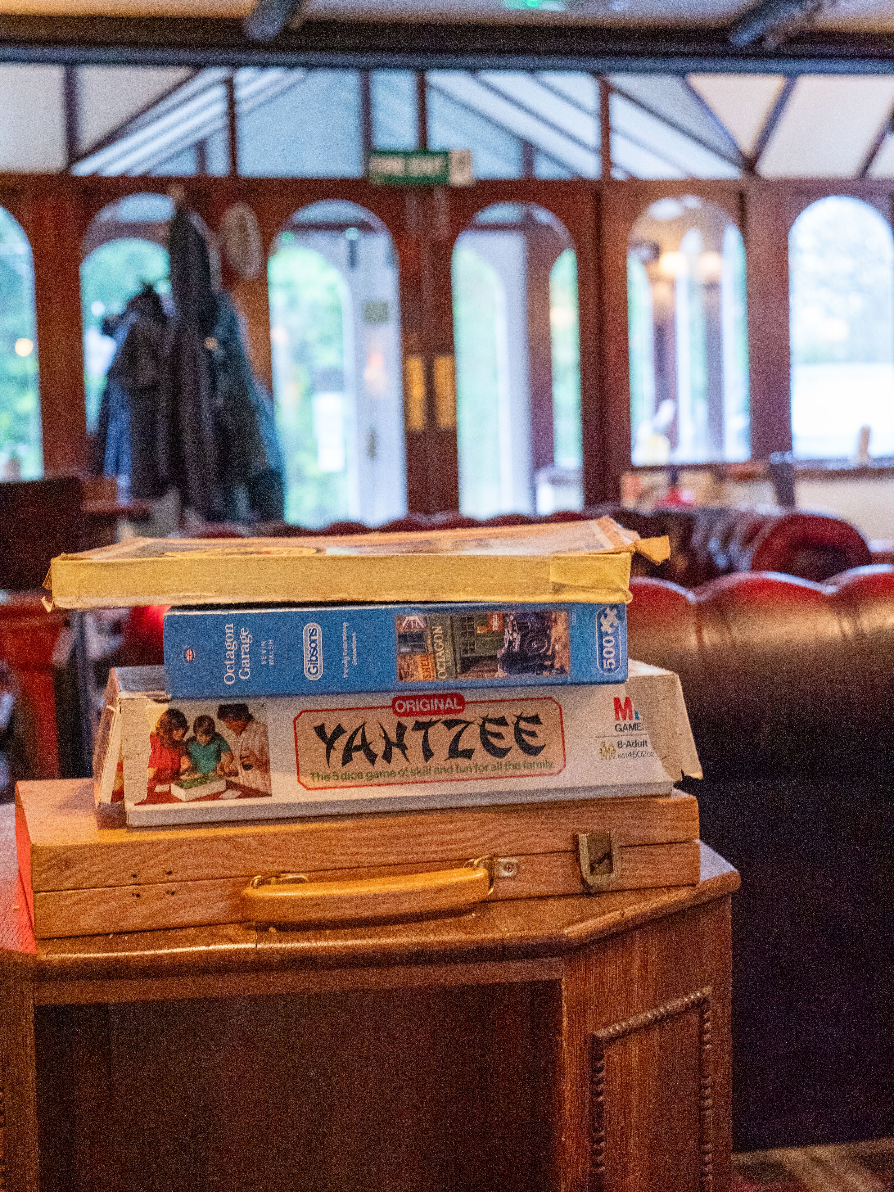A stack of board games on a wooden table in a room with a high ceiling and arched windows.