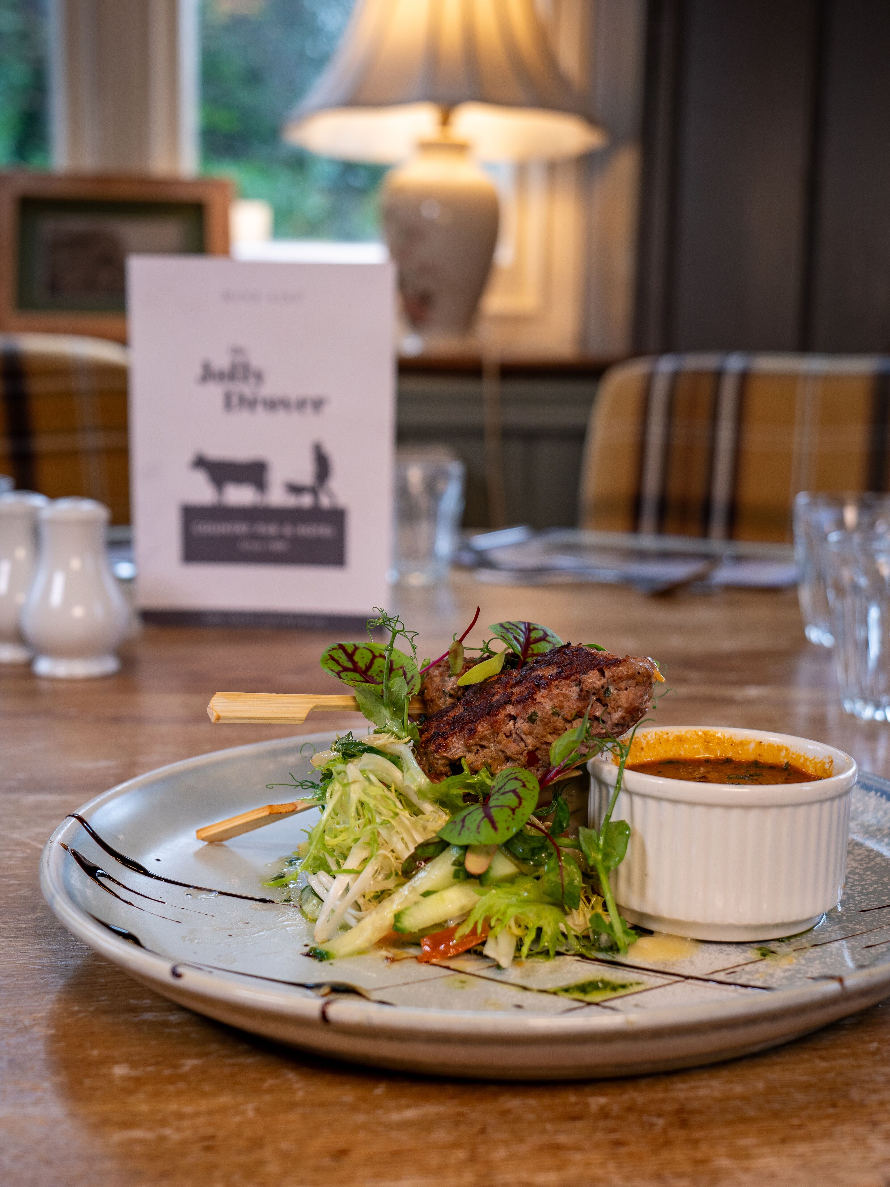 A plate of food with a meat dish, greens, and a small bowl of sauce, set on a table with a menu and salt shakers in the background.