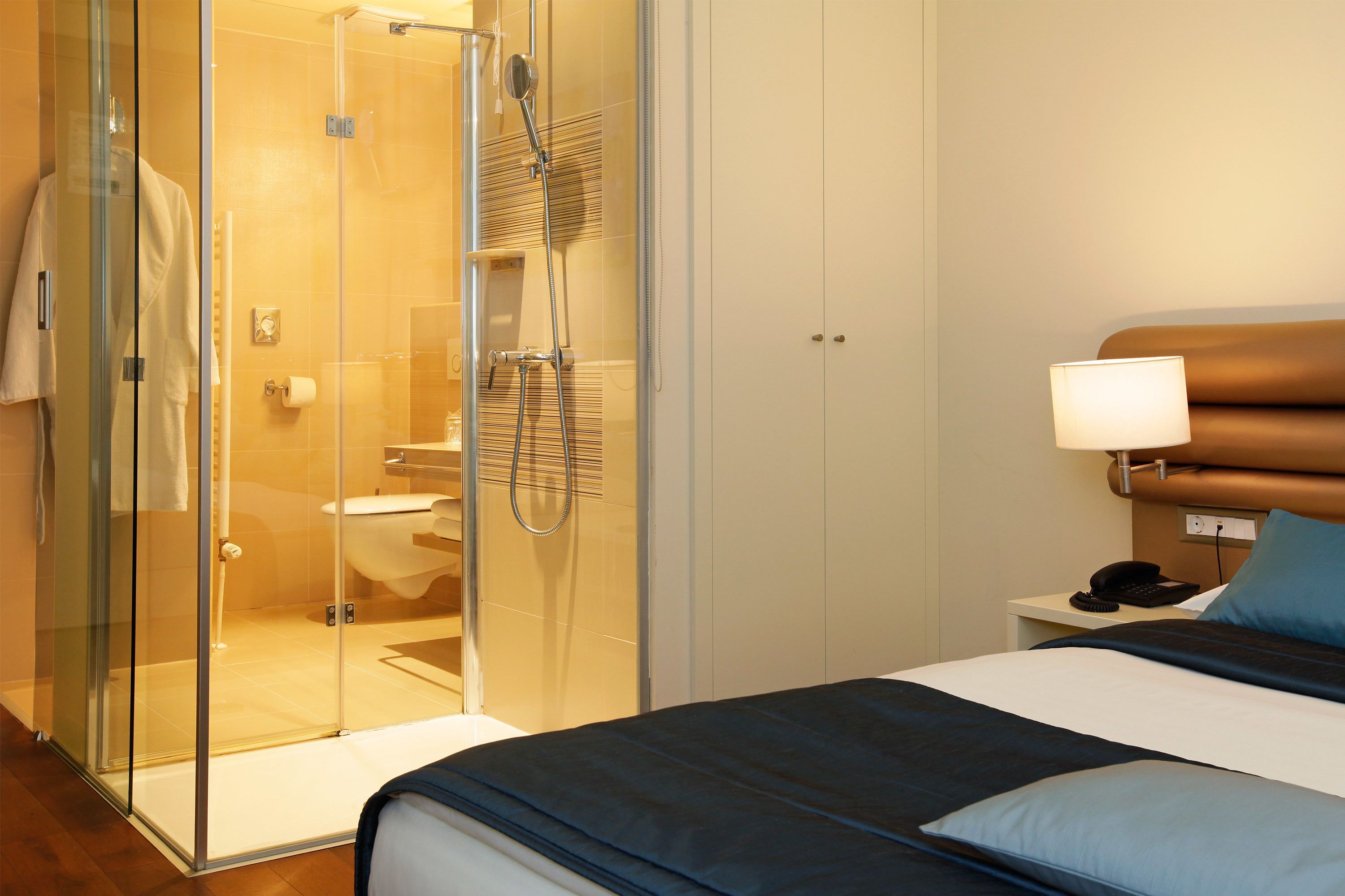 Camera d'albergo con cabina doccia in vetro, un letto con testiera, un comodino con una lampada e un tavolino con il telefono.