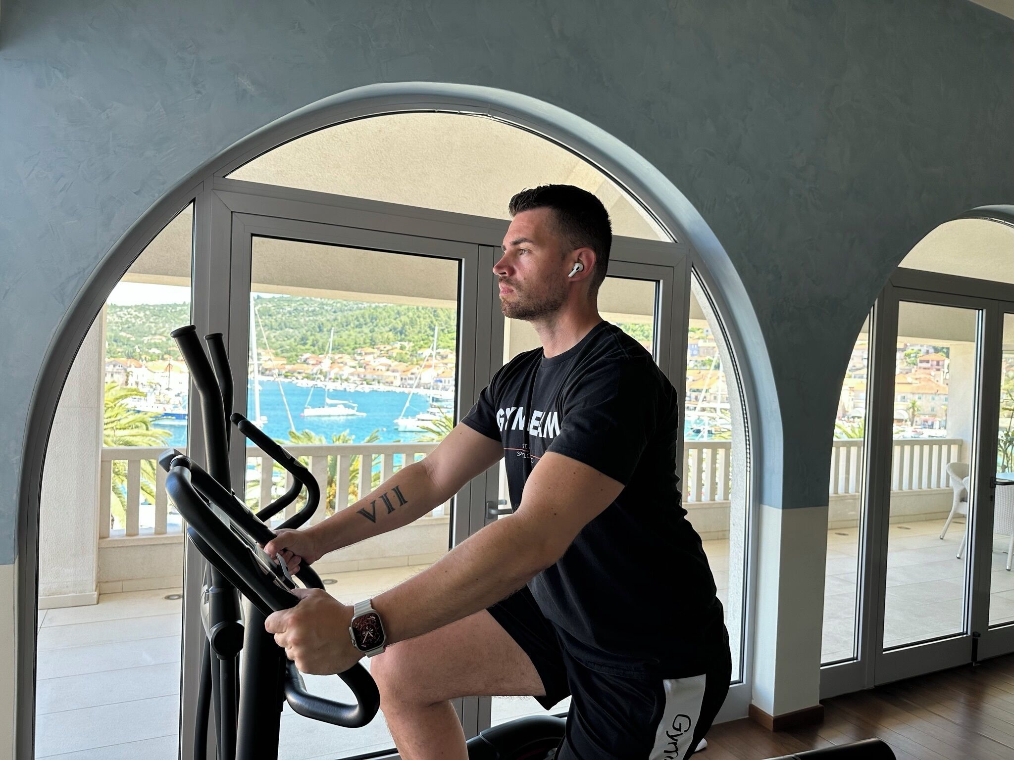 Een man sport op een hometrainer in een kamer met grote ramen en uitzicht op een kustplaats.