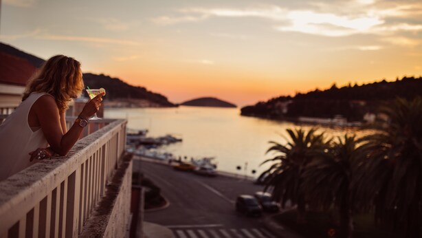 Een vrouw zit op een balkon met uitzicht op een wateroppervlak met boten en een zonsondergang.