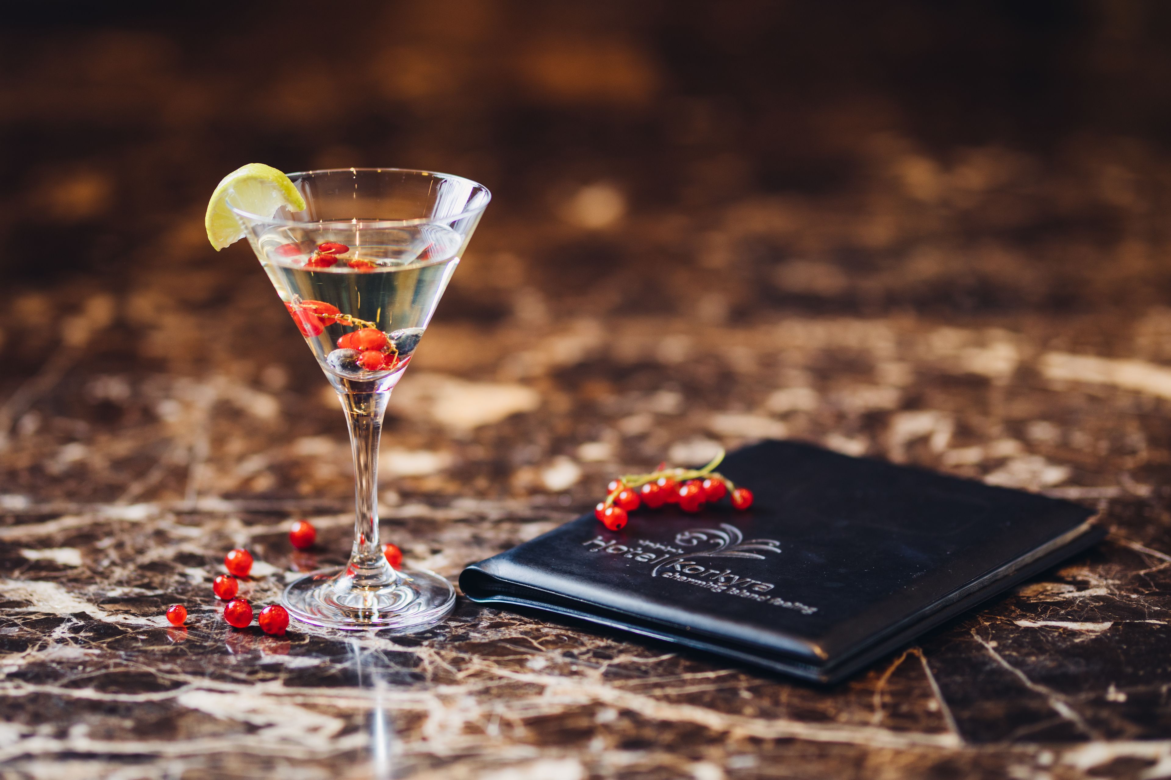 A martini glass with a lime wedge and red berries on a marble surface, next to a black notebook.