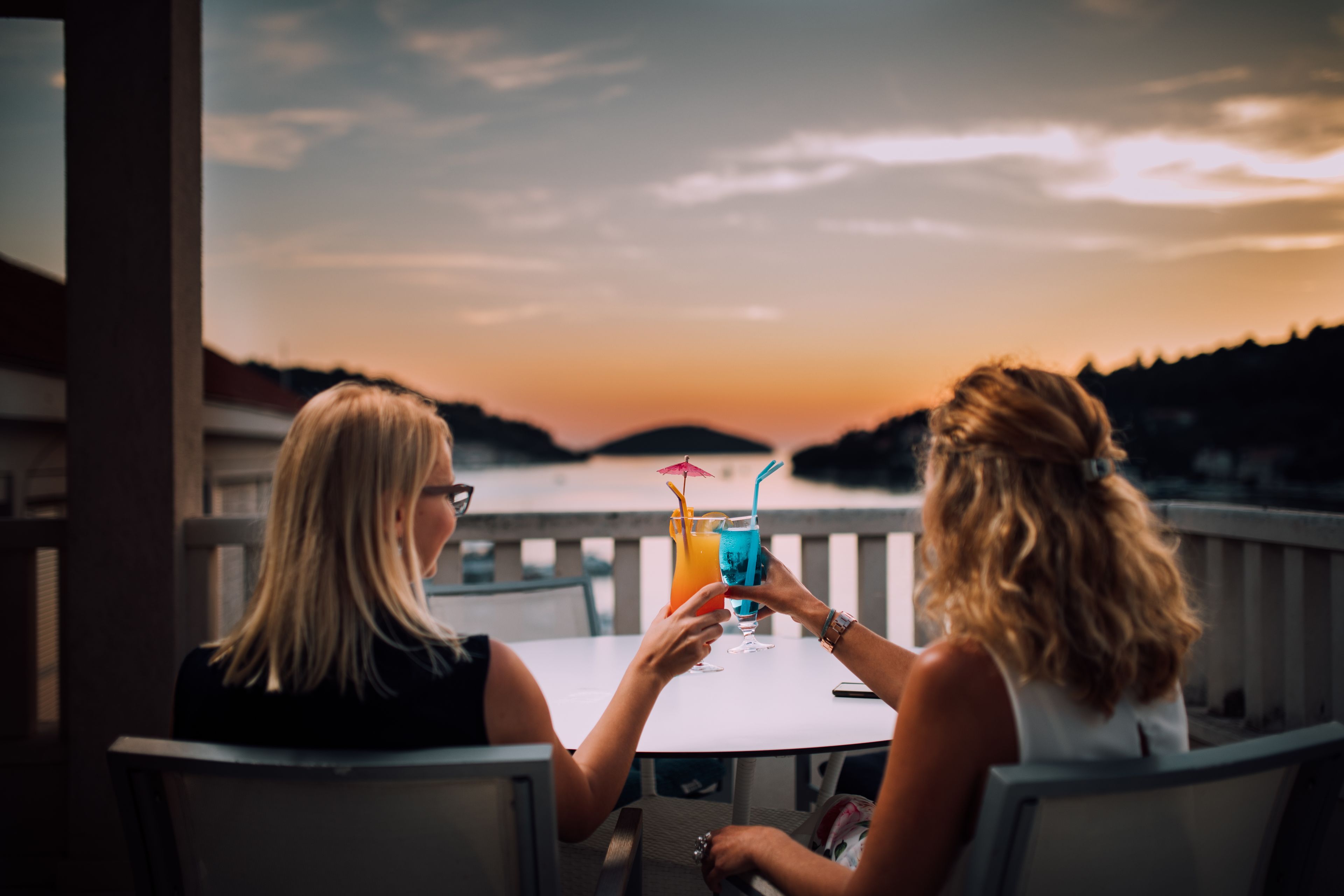 Two people sitting at a table with drinks, overlooking a scenic view of hills and a sunset.