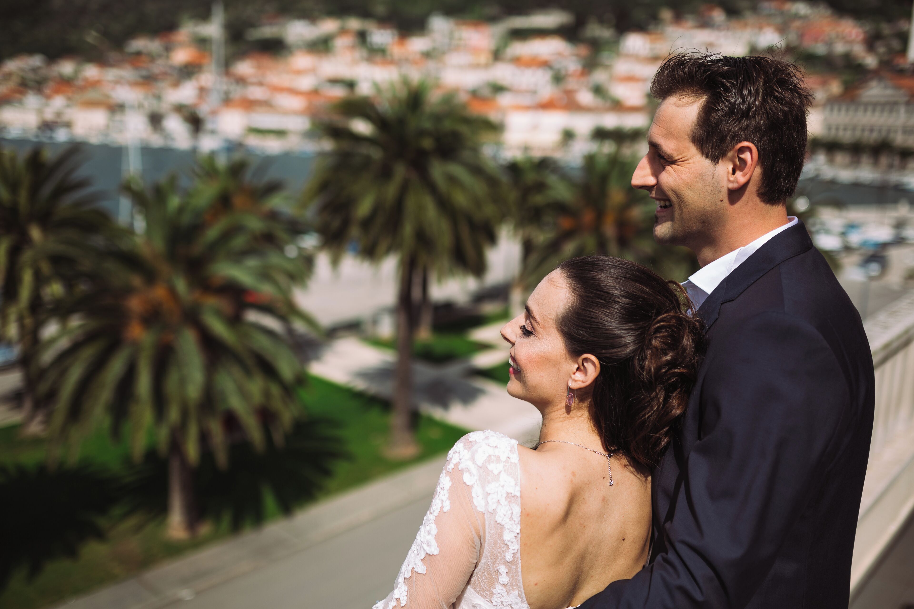 Una pareja se abraza en un balcón con vista a una ciudad con palmeras y edificios.