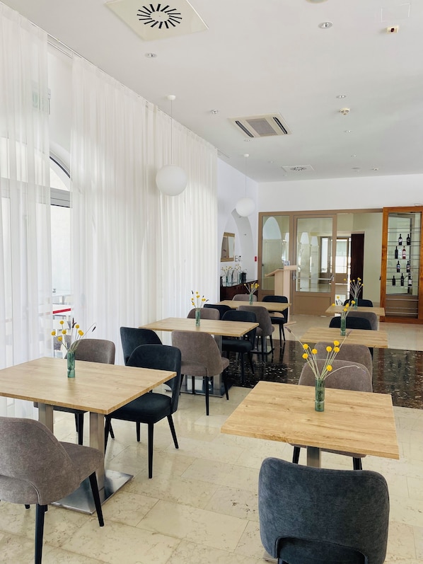 A modern dining area with wooden tables and chairs, white curtains, and a view of the kitchen in the background. - Hotel Korkyra (Vela Luka)
