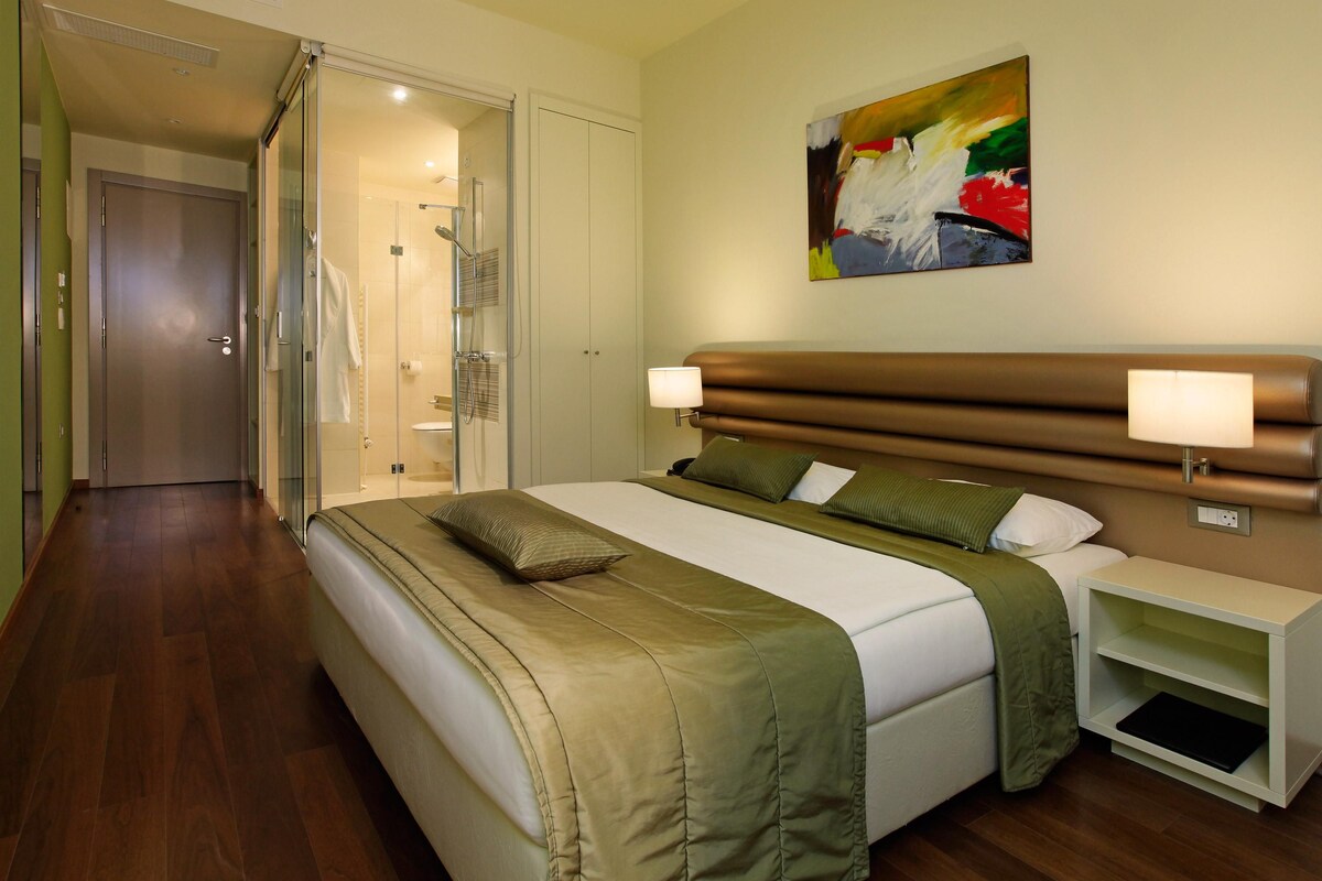 A hotel room with a large bed, a bedside table, a shower area, and a painting on the wall.