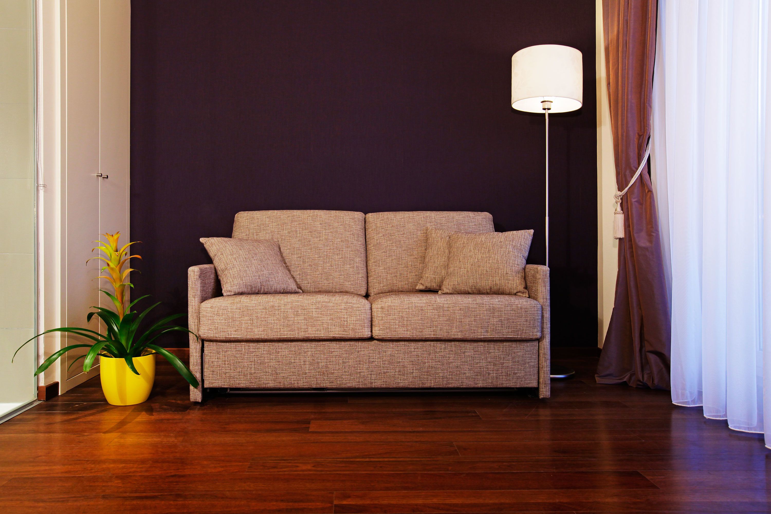 Un canapé beige avec deux coussins, une plante en pot jaune, une lampe sur pied et un mur foncé.