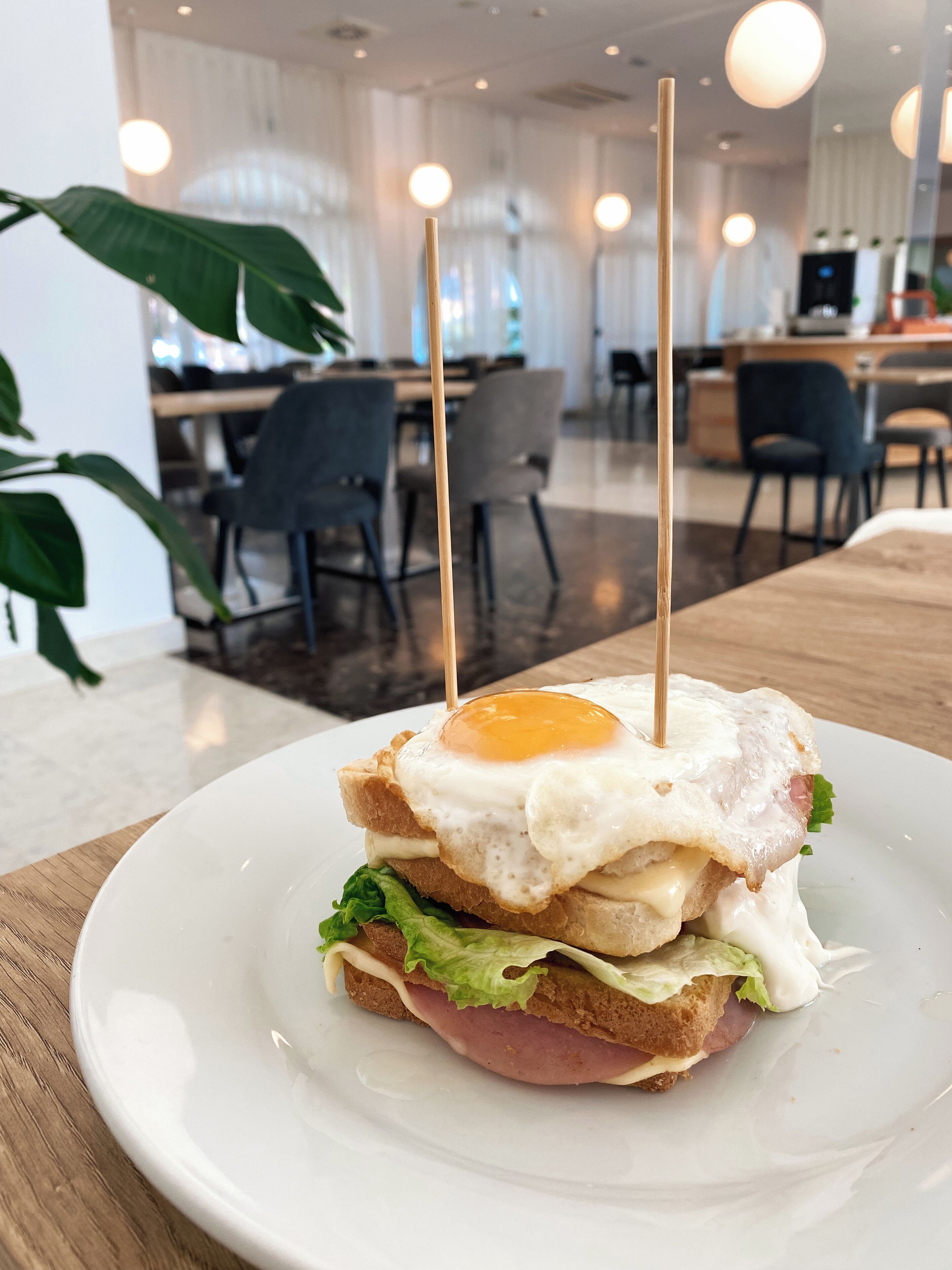 A sandwich with a fried egg on a white plate, set on a wooden table in a modern dining area with chairs and tables.