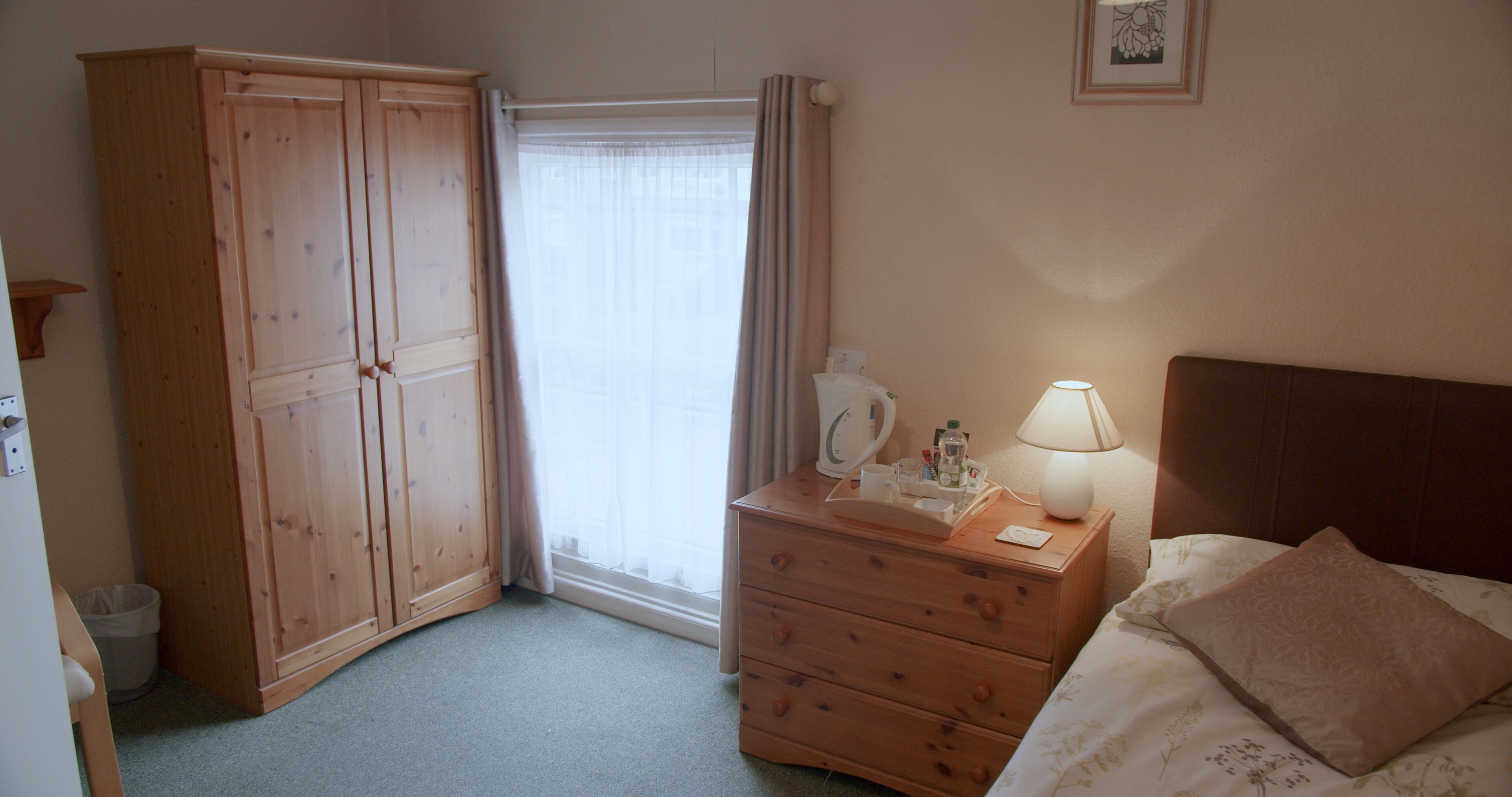 A bedroom with a wooden wardrobe, a bedside table with a lamp, a bed with a floral bedspread, and a window with curtains.