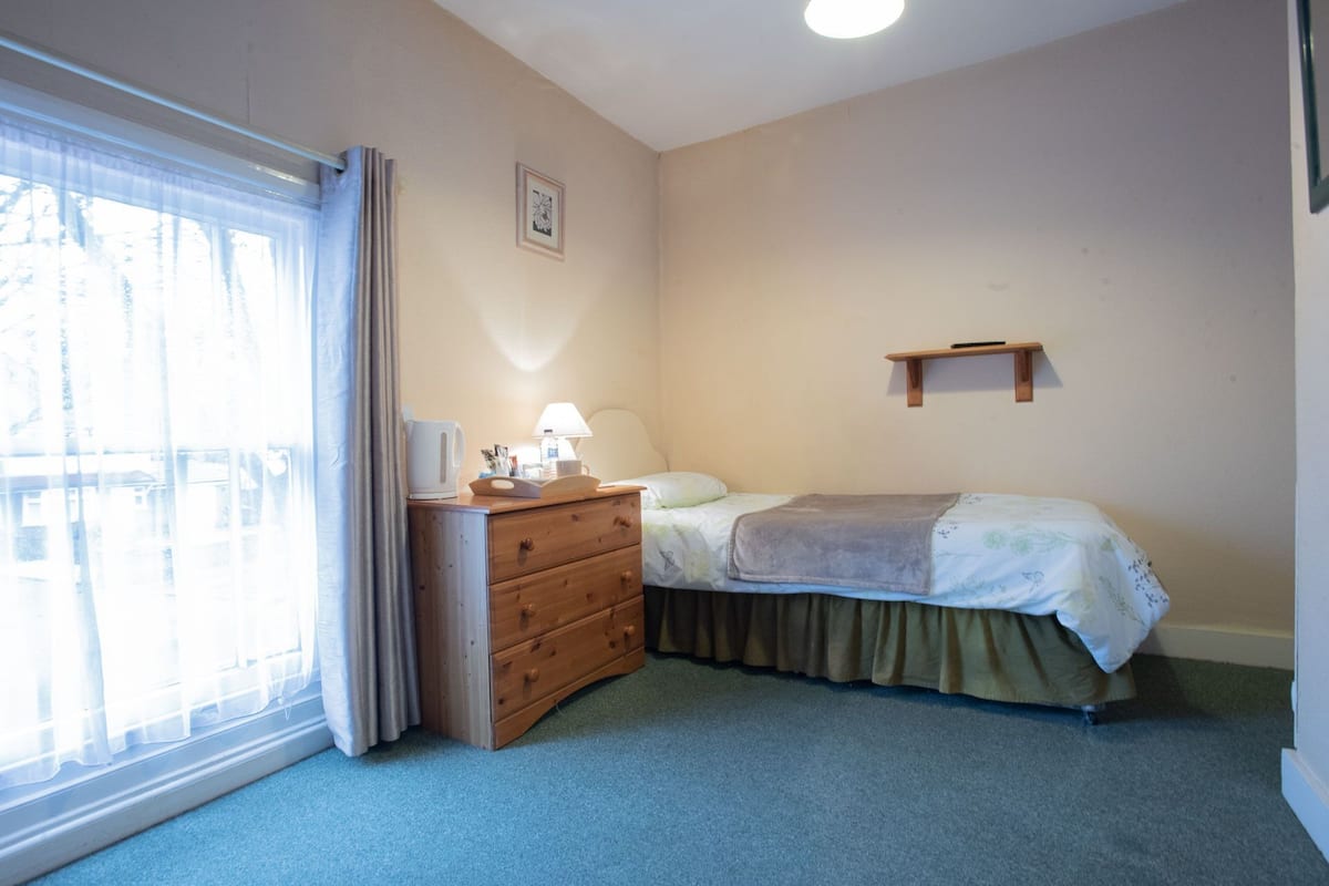 A bedroom with a bed, a dresser, a bedside table with a lamp, a window with curtains, and a wall-mounted shelf.