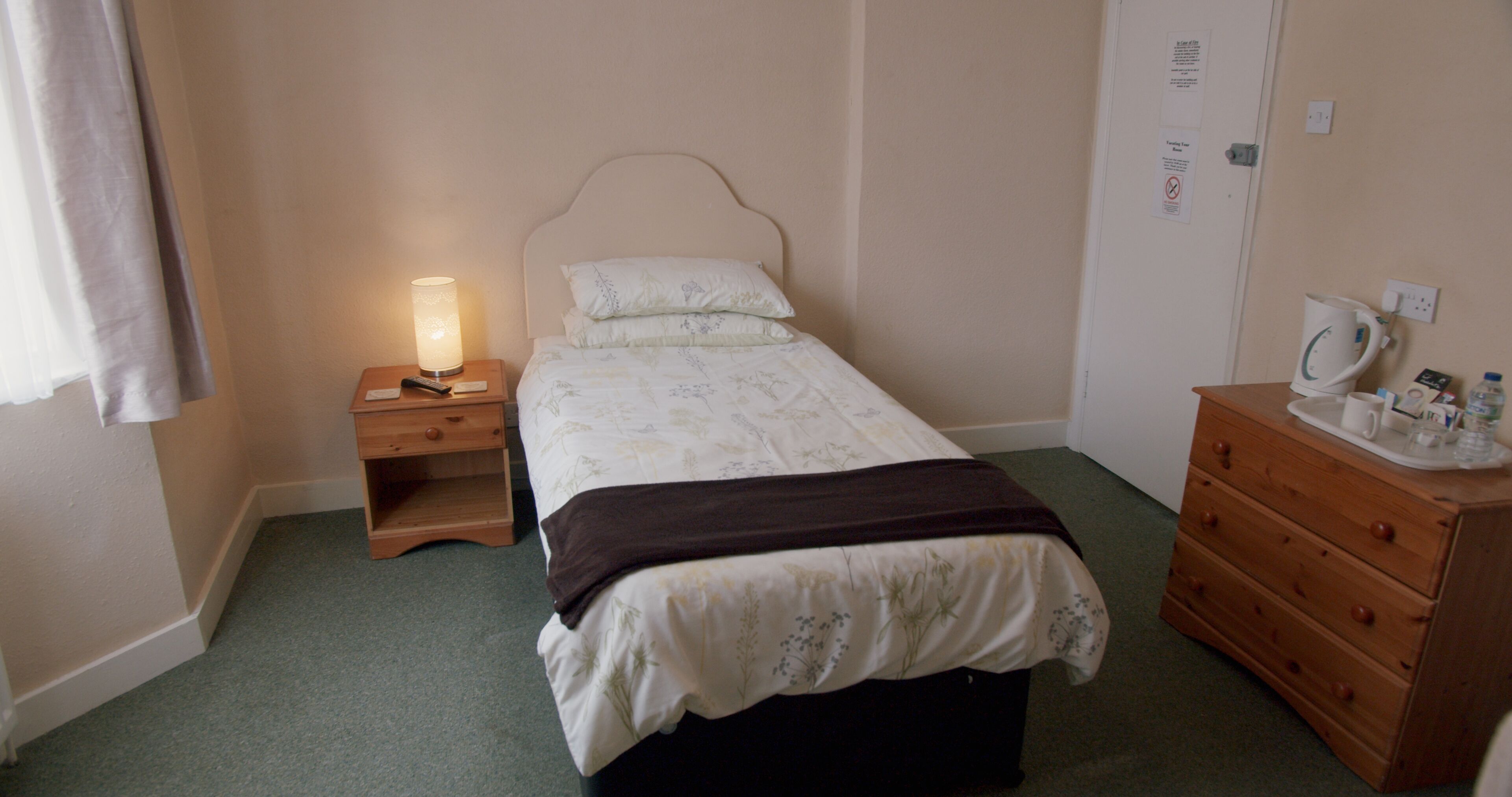 A bedroom with a bed, bedside table, lamp, dresser, and a door with a sign.