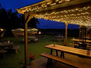 An outdoor patio area with wooden tables and benches, string lights, and a pergola structure. - The Allanton Inn (Duns)