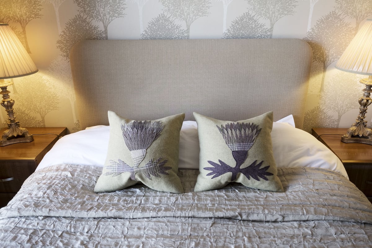 A neatly made bed with a textured gray bedspread, two decorative pillows with a leaf pattern, and a headboard against a wall with a tree pattern.