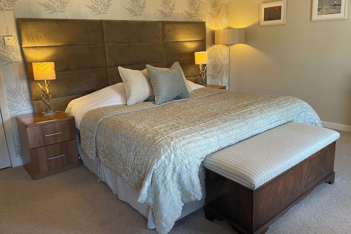 A bedroom with a large bed, two bedside tables with lamps, a bench, and patterned wallpaper.