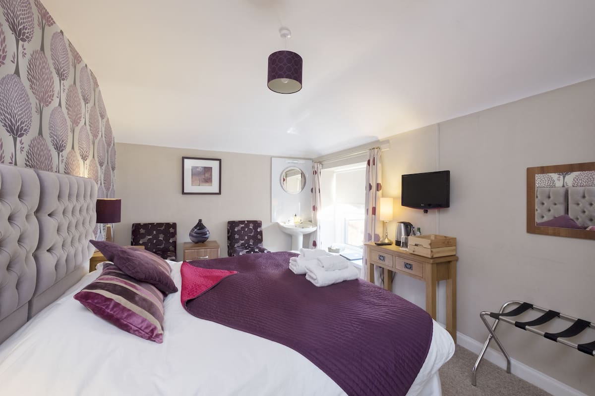 A neatly arranged bedroom with a large bed, a bedside table, a mirror, a TV, and a window with curtains.