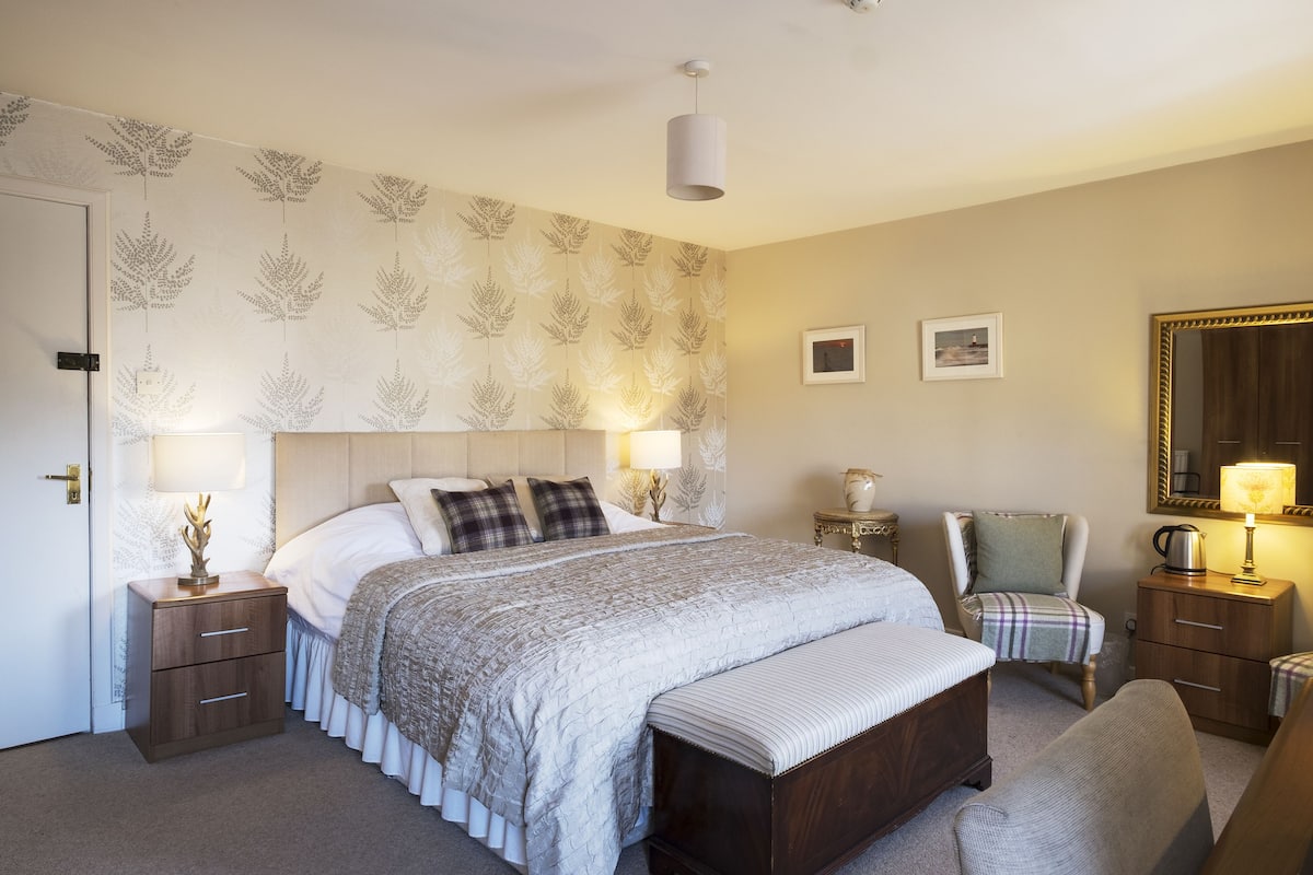 A neatly arranged bedroom with a large bed, bedside tables, a chair, a bench, and a mirror.