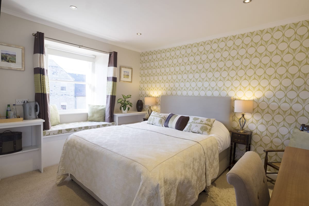 A bedroom with a bed, bedside lamps, a window with curtains, a bedside plant, and a small table with a kettle.