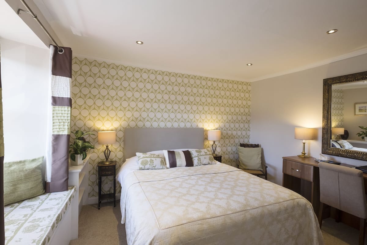 A hotel room with a bed, bedside tables, a desk, a mirror, and patterned wallpaper.