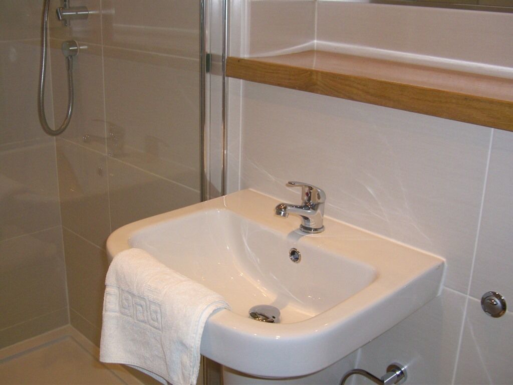 Un bagno con un lavandino bianco, una doccia e uno scaffale in legno sopra il lavandino.
