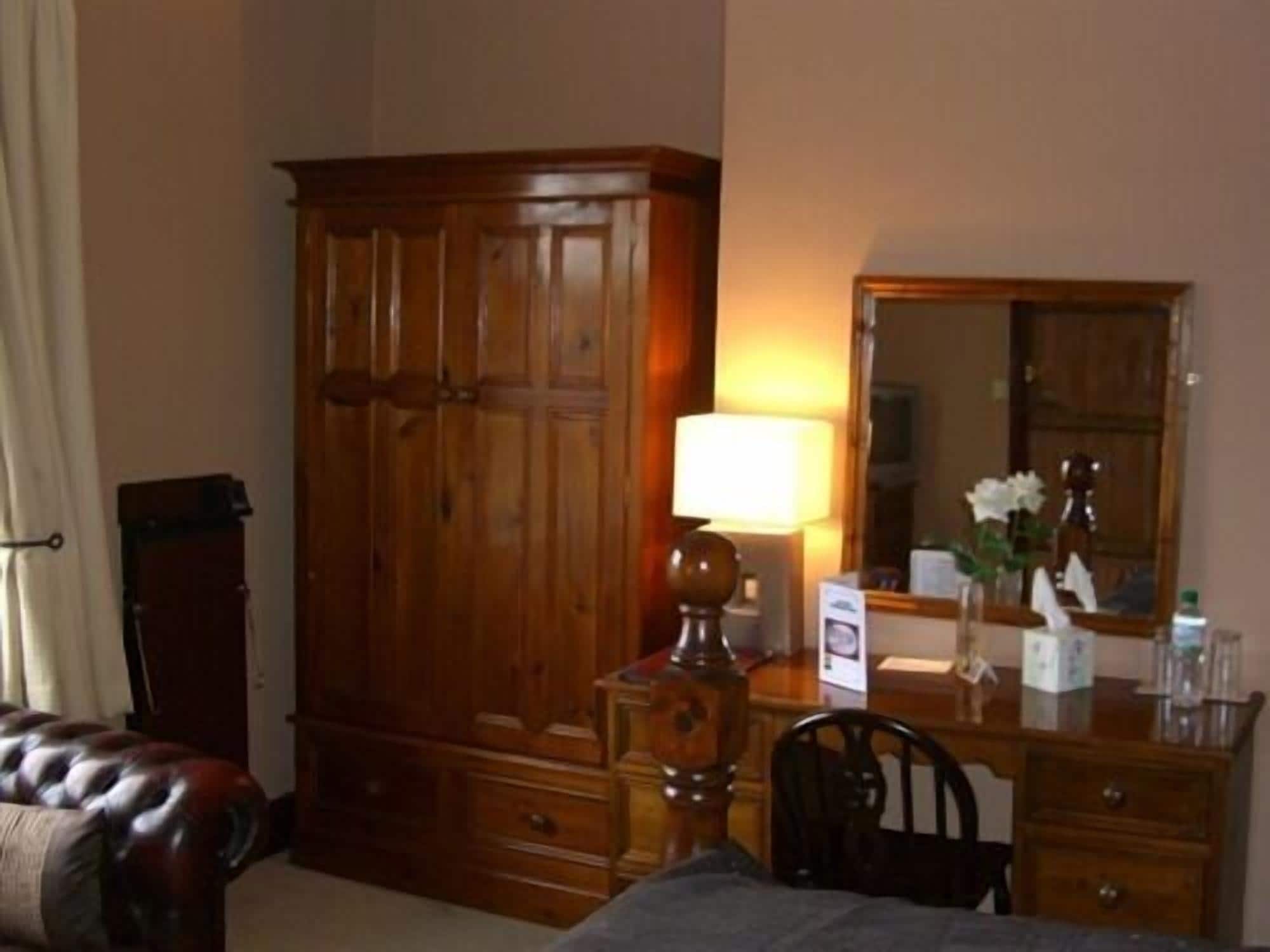 A hotel room with a wooden wardrobe, a bedside table with a lamp, a mirror, and a chair.
