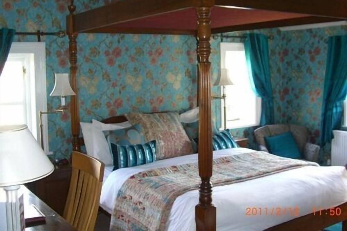 A four-poster bed with a floral-patterned bedspread, a wooden headboard, and a window with curtains.