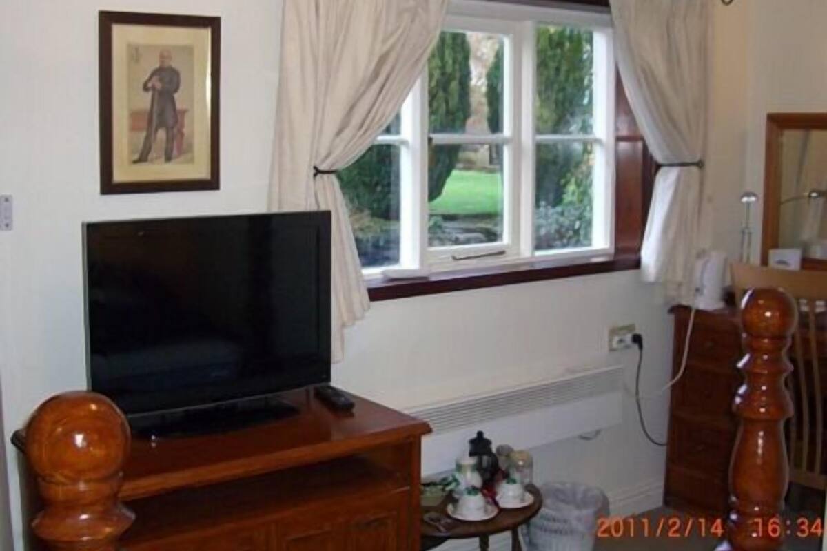 A room with a television on a wooden stand, a window with curtains, a small table with items, and a mirror.