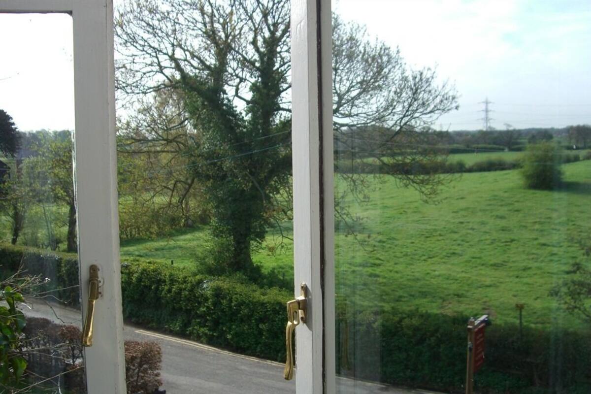 A view through a window with a white frame and brass handles, overlooking a rural road and green fields.