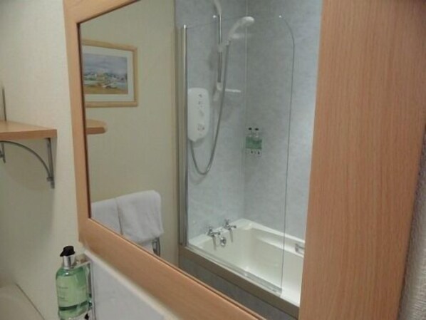 Un baño con ducha de vidrio, una bañera blanca, un estante de madera con un cuadro enmarcado y una botella de champú.