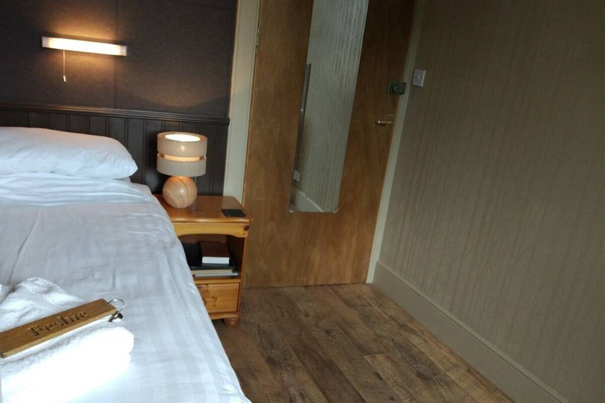 A hotel room with a bed, a bedside table with a lamp, a wooden nightstand, and a door with a towel hanging on it.