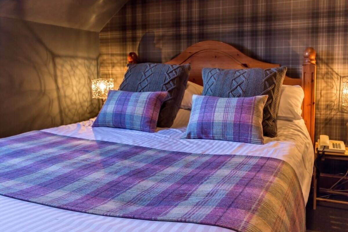 A cozy bedroom with a plaid bedspread, a wooden headboard, and a nightstand with a lamp and phone.