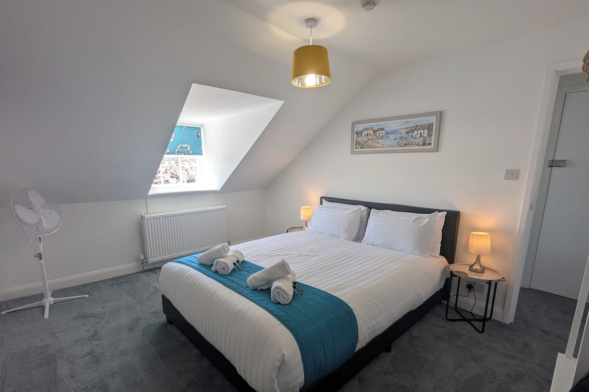 A bedroom with a bed, a standing fan, a radiator, a window with a view, and a painting on the wall.