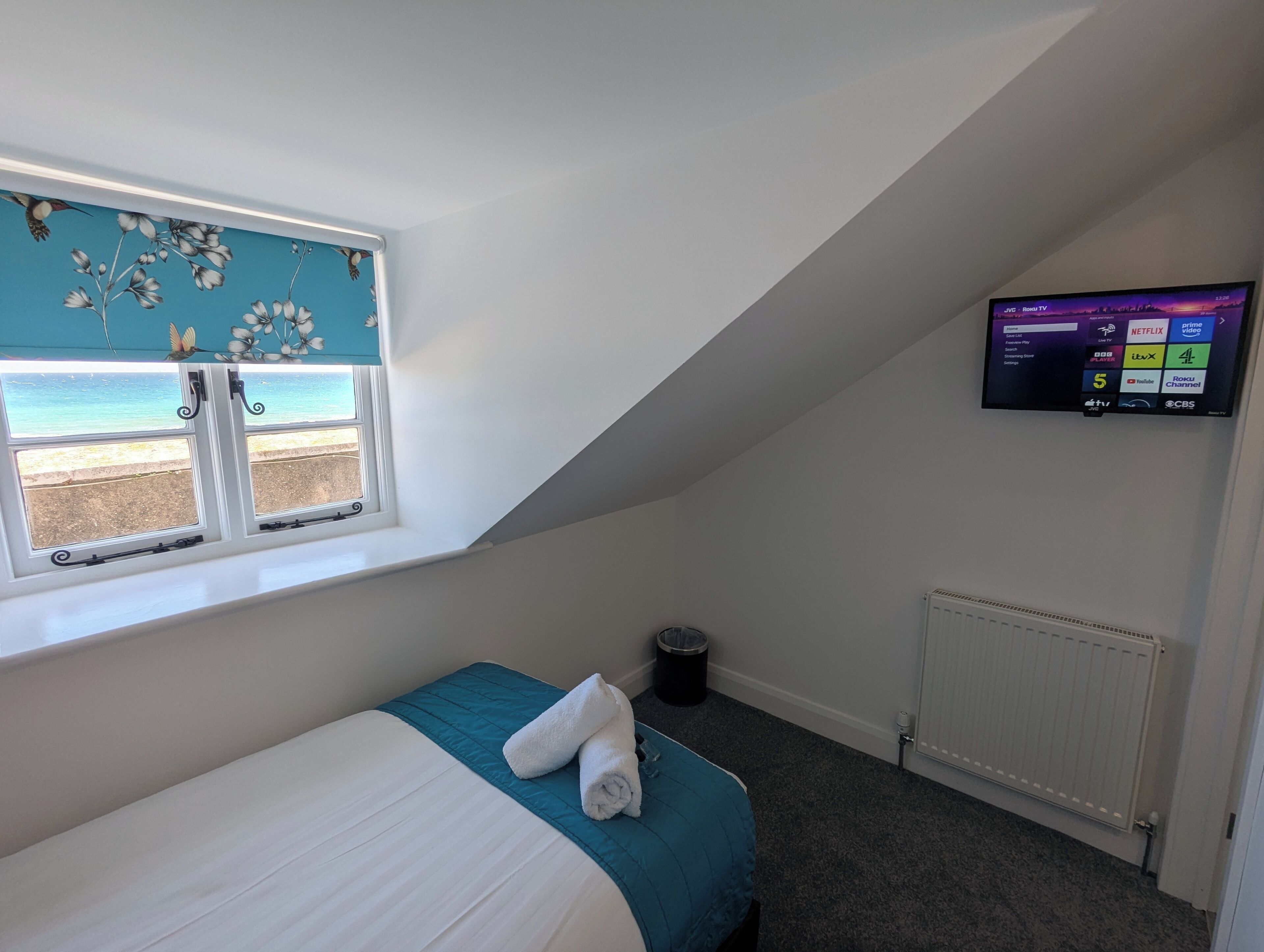 A bedroom with a bed, a window with a view of the beach, a television on the wall, and a radiator.