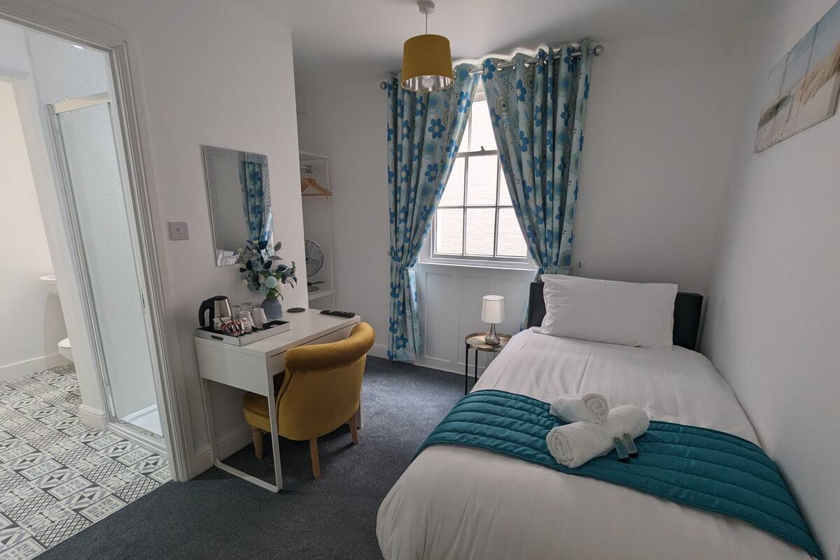 A hotel room with a bed, a desk with a chair, a small table with a lamp, and a window with curtains.