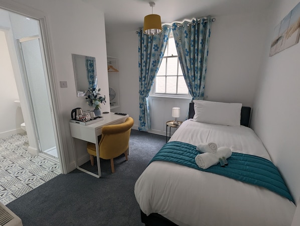 Habitación de hotel con una cama, un escritorio con silla, una mesita de noche con lámpara y una ventana con cortinas.