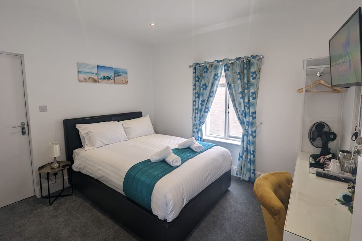 A neatly made bed with white and teal bedding, a small bedside table with a lamp, a window with blue floral curtains, and a desk with a fan and personal items.