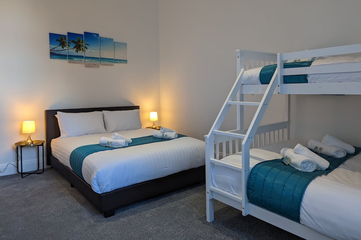 A hotel room with a bunk bed, a single bed, and a nightstand with a lamp. There are towels on the beds and a painting on the wall.