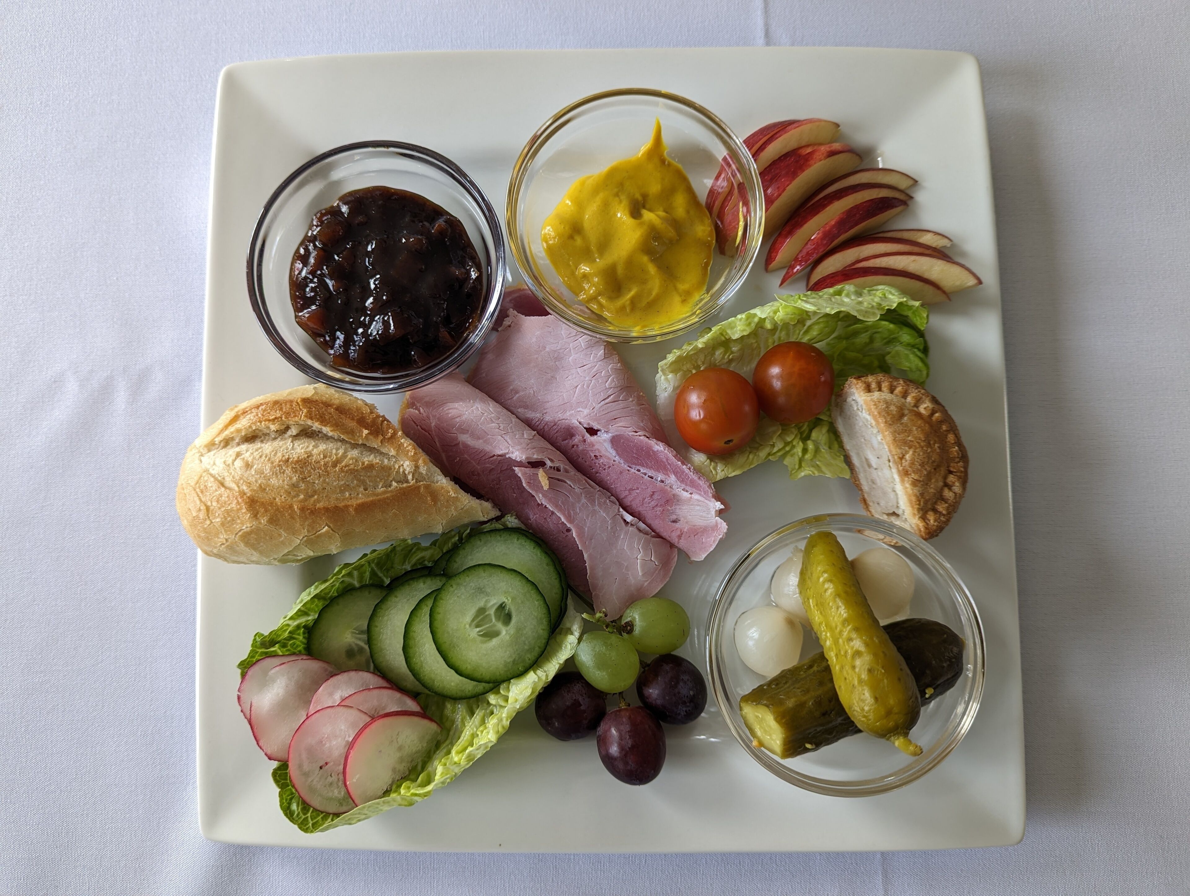 A white rectangular plate with various food items including ham, bread, lettuce, radishes, grapes, pickles, and condiments.