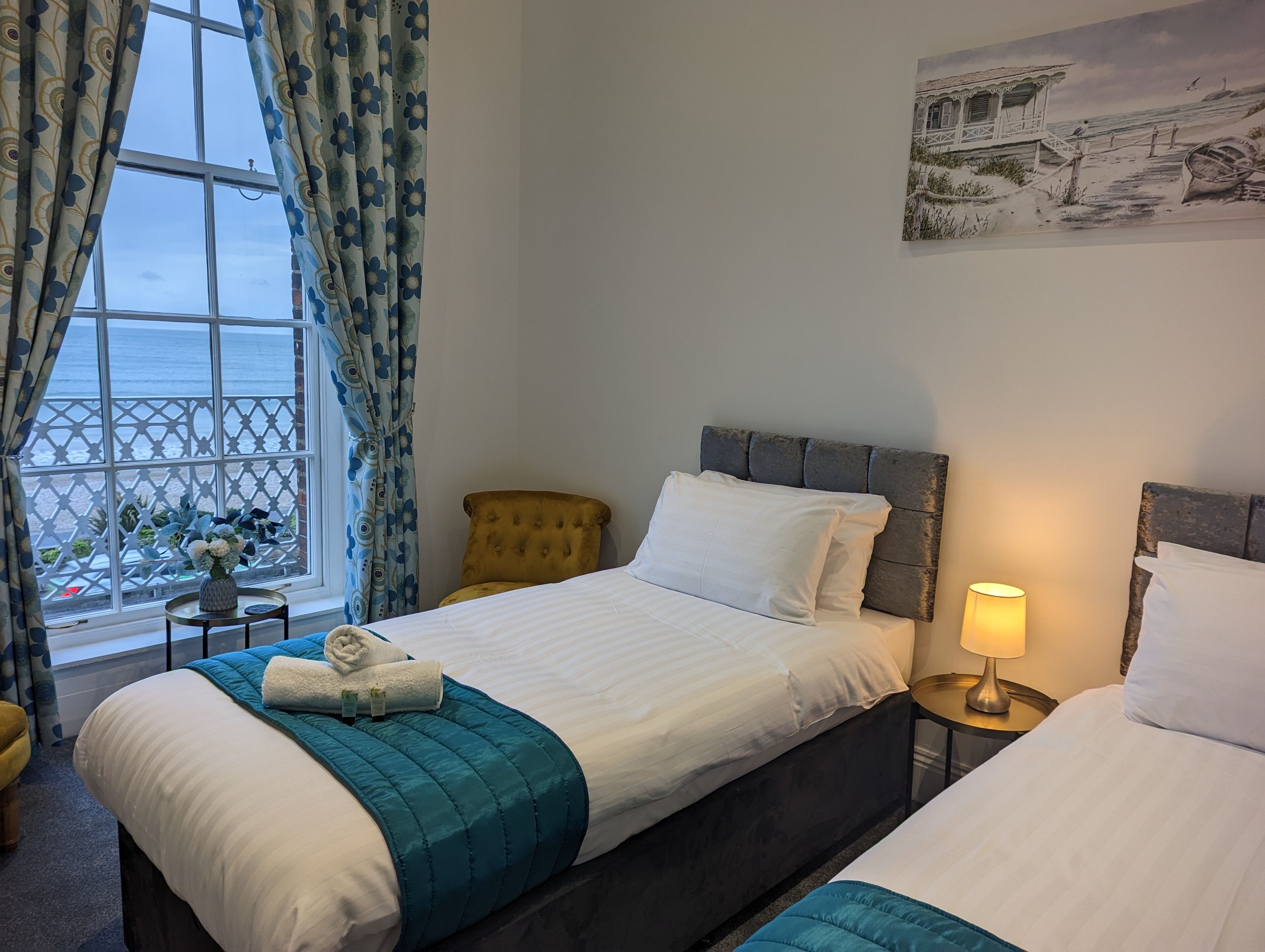 A hotel room with two beds, a window with curtains, a small table with a vase, and a painting on the wall.