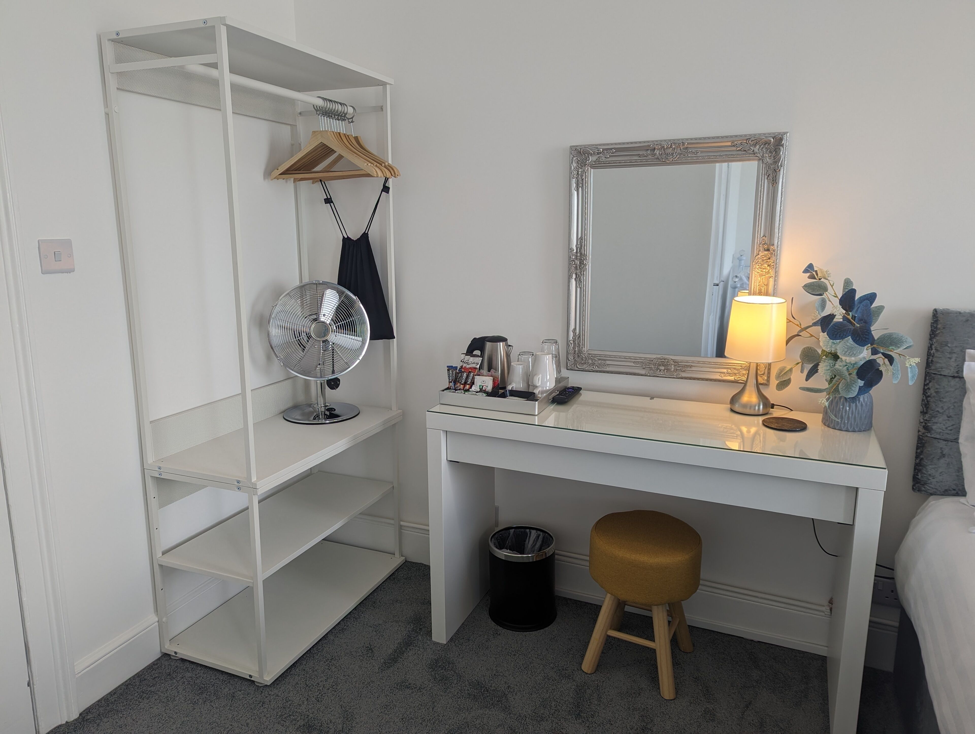 Un bureau blanc avec un miroir, un ventilateur, un petit tabouret et une poubelle.