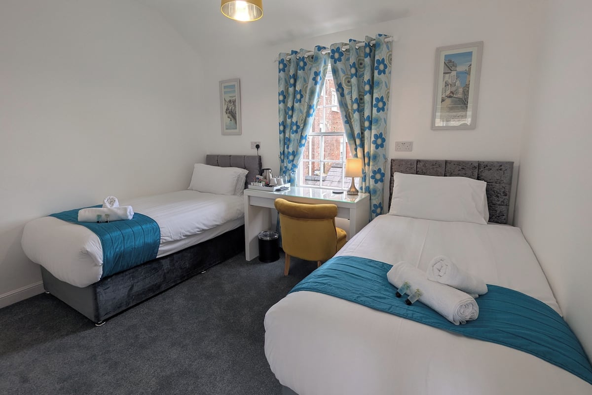 A hotel room with two single beds, a desk with a chair, a lamp, and a window with blue floral curtains.