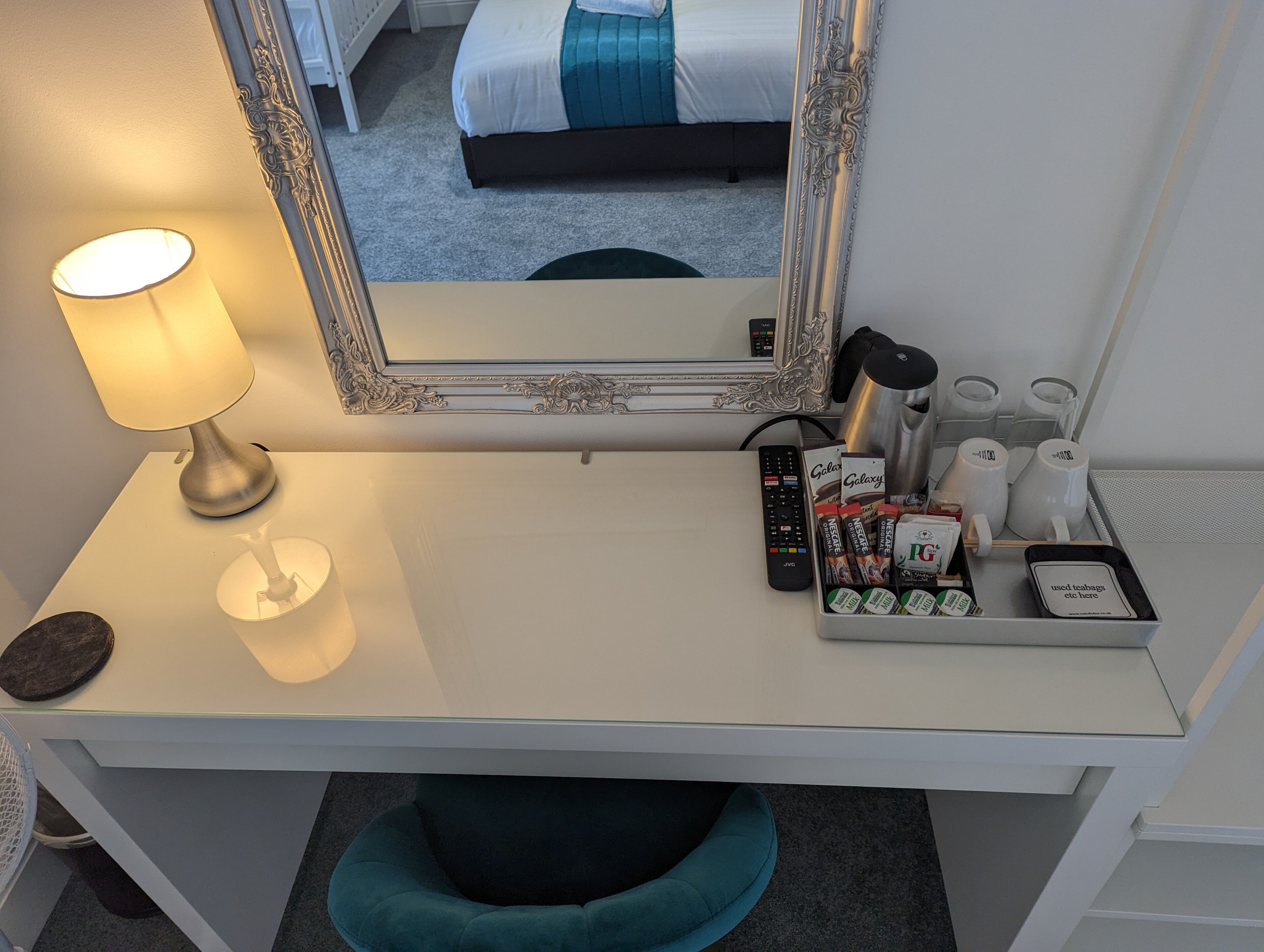 A hotel room desk with a lamp, a mirror, a remote control, and a tray with toiletries.